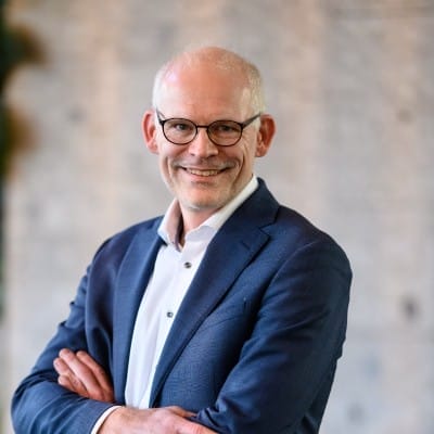 Een man in een blauw pak en wit overhemd staat met zijn armen over elkaar en glimlacht zelfverzekerd voor een zacht vervaagde achtergrond die zijn expertise in cyberbeveiliging en beveiligingsmonitoring weerspiegelt.